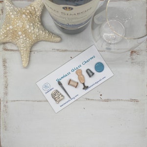 May include: A collection of stemless glass charms with sewing-themed designs, including a needle, thread spool, dress form, thimble, and a blue charm. The charms are displayed on a white card with the text "Stemless Glass Charms". A starfish, wine bottle, and glass are also present.