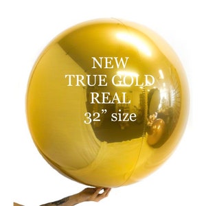 Puede incluir: Globo grande y redondo de color dorado con superficie reflectante. El globo está sostenido por una mano y presenta el texto "NEW TRUE GOLD REAL 32" size" en blanco. El globo tiene aproximadamente 81 cm de diámetro.