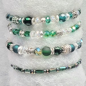 May include: A set of five green and silver beaded bracelets. The bracelets are made with various sizes and shapes of green beads, including round, square, and oval beads. The beads are interspersed with silver spacers and charms.