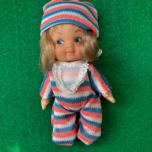 Può includere: Bambola vintage con tuta a righe e cappello abbinato nei toni del rosso, bianco e blu. La bambola ha i capelli biondi e gli occhi azzurri. L'outfit include un bavaglino bianco.