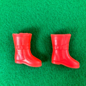 May include: A pair of bright red plastic boots, likely for a doll or toy, are displayed against a green background. The boots have a classic rain boot design with a buckle detail. The boots are small, suggesting they are for a child's toy.