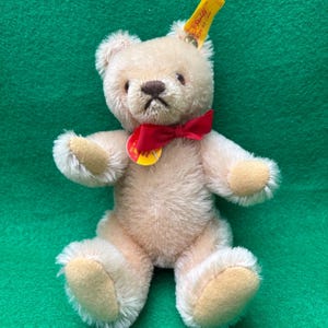 May include: A small, plush teddy bear with light brown fur, a red bow tie, and a yellow tag. The bear has a stitched brown nose and mouth, and tan-colored paw pads. The bear is sitting on a green surface.
