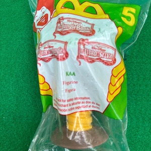 May include: Sealed plastic bag containing a yellow and brown figurine from "The Jungle Book." The bag has a green and white design with the number 5. Text includes "Figurine" and "KAA."