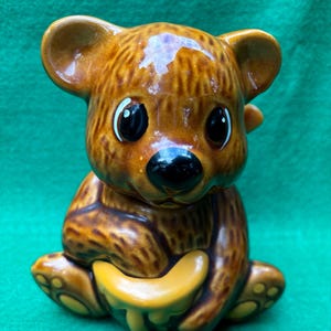 May include: A brown ceramic bear figurine holding a honey pot. The bear has a cute, cartoon-like face with big eyes and a black nose. The honey pot is yellow and dripping with honey.