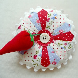 Könnte beinhalten: Ein weißes Nadelkissen mit einem roten und blauen Blumenmuster und einer roten Stoff-Erdbeere oben drauf. Das Nadelkissen hat einen gewellten Rand.