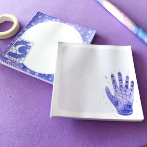 May include: A stack of purple and white notepads with a moon and stars design. The top notepad features a hand with astrological symbols and lines.