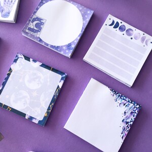 May include: Four different square sticky note pads with celestial and moon themed designs. The note pads are on a purple surface. The designs include a full moon, a crescent moon, a moon phase cycle, and a crystal cluster.