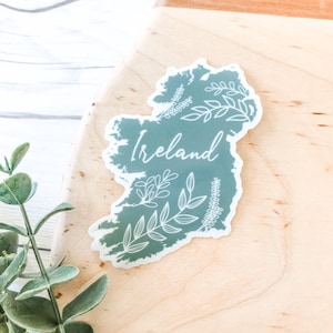 May include: A green sticker with a white outline of the island of Ireland. The sticker has a white floral design and the word "Ireland" written in white cursive.