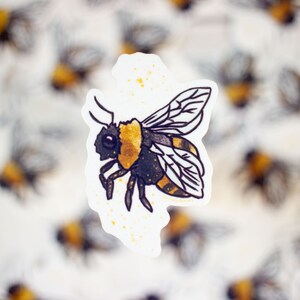 May include: A black and yellow bumblebee sticker with gold glitter accents. The sticker is on a white background with a blurred image of other bumblebee stickers in the background.