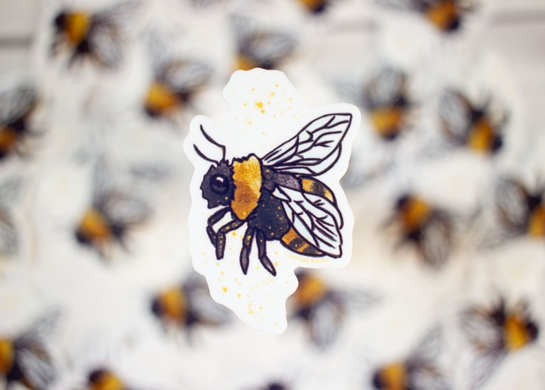 May include: A black and yellow bumblebee sticker with gold glitter accents. The sticker is on a white background with a blurred image of other bumblebee stickers in the background.