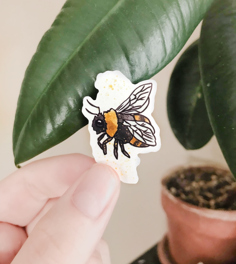 May include: A yellow and black bumblebee sticker with gold glitter accents. The sticker is on a white background.