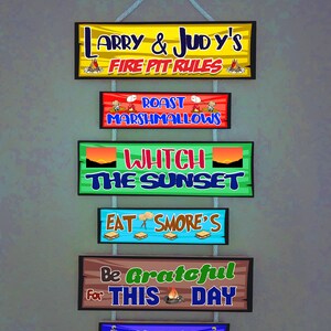 May include: A hanging wooden sign set with six rectangular panels connected by rope. The text reads: LARRY & JUDY'S FIRE PIT RULES, ROAST MARSHMALLOWS, WHTCH THE SUNSET, EAT SMORE'S, Be Grateful for THIS DAY, and Friends & Family are always welcome.