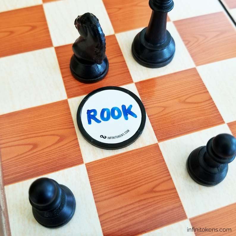 May include: A chessboard with black and white squares, displaying black chess pieces and a circular token. The token has the word "ROOK" in blue watercolour lettering and the website address "infinitokens.com".