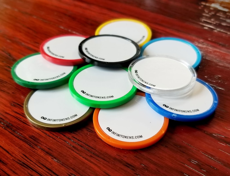 May include: A selection of circular tokens with white centres and the text INFINITOKENS.COM. The tokens have coloured edges in red, green, black, blue, orange, yellow, and clear. The tokens are arranged on a wooden surface.