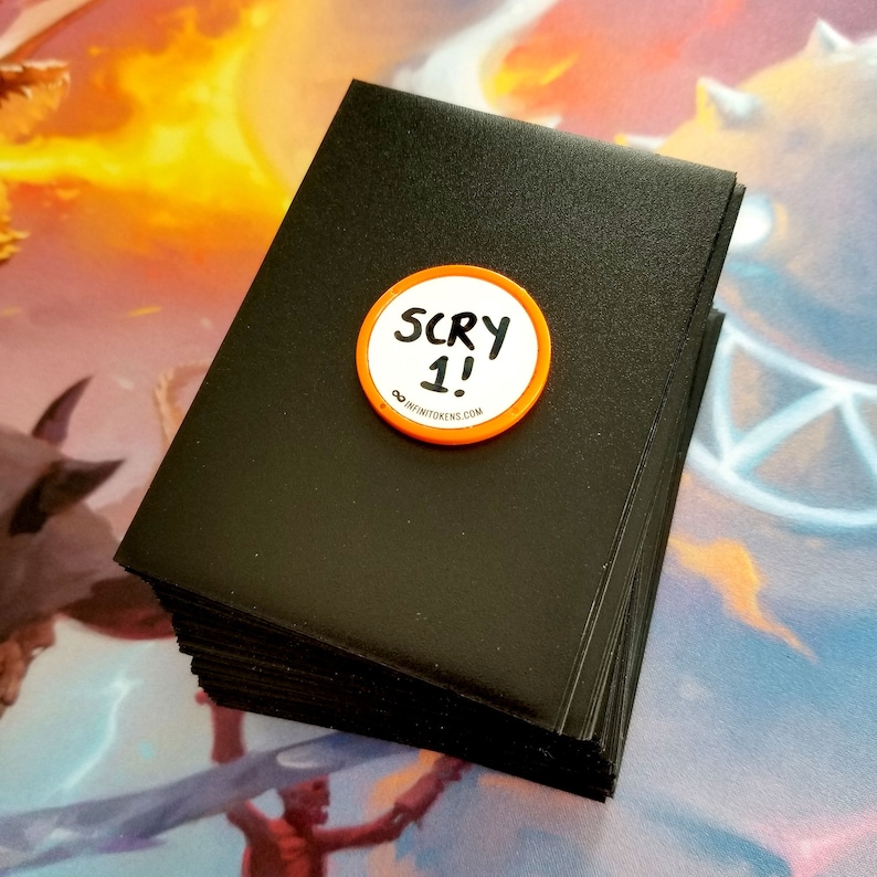 May include: A pile of black card sleeves with a circular orange and white token on top. The token reads "SCRY 1!" and "&infin;infinitetokens.com". The card sleeves are likely for trading card games.