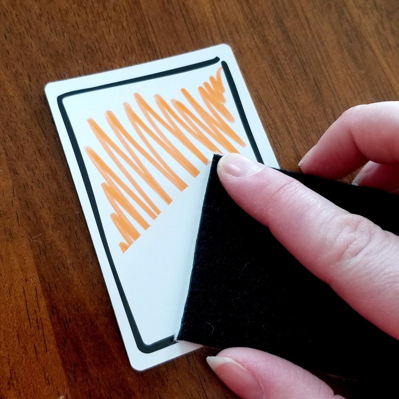May include: A white card with a black border and orange lines drawn inside. A hand is holding a black eraser over the card.