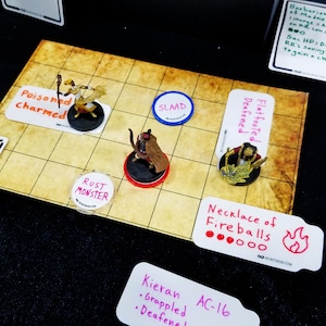 May include: A tabletop role-playing game setup with miniature figures on a grid map. The scene includes character cards, status markers, and a "Necklace of Fireballs" card. The figures are on round bases, and the cards have handwritten text.