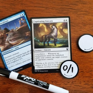 May include: Two Magic: The Gathering cards, "Captivating Unicorn" and "Morphosis", with a black dry-erase marker and two white tokens with "0/1" printed on them. The cards are on a wooden surface.