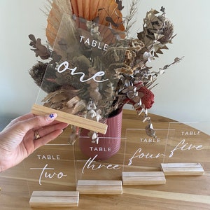 May include: Five clear acrylic table number signs with wooden bases. The numbers are written in white cursive font. The numbers are one, two, three, four, and five.
