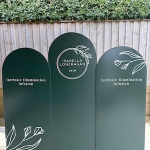 May include: Three dark green arched panels with white text and floral designs. The centre panel reads "Isabella Loneragan Skin". The left and right panels read "Intrinsic Illumination Infusion".