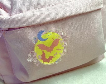 Pode incluir: Uma mochila rosa com um alfinete decorativo com uma lua amarela, morcegos rosa e uma lua crescente azul. O alfinete tem uma borda decorativa transparente. O texto "SULKY BLACK HEART" é visível na parte inferior.
