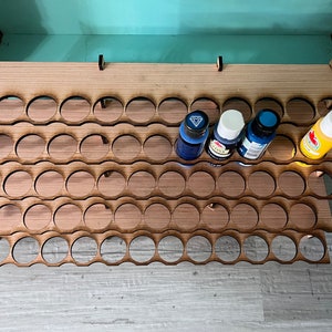 May include: A wooden paint rack with three tiers, each tier has multiple circular cutouts to hold paint bottles. The rack is made of light brown wood and has a natural wood finish. Four paint bottles are visible, two blue, one white, and one yellow.