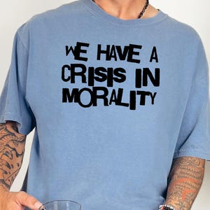 Op de afbeelding: Lichtblauw t-shirt met de tekst "WE HAVE A CRISIS IN MORALITY" in dikke zwarte letters. Het shirt is effen van kleur en heeft korte mouwen. Het shirt wordt door een persoon gedragen.