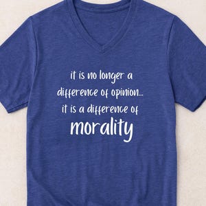 Op de afbeelding: Een blauw V-hals T-shirt met de witte tekst: "het is niet langer een verschil van mening... het is een verschil van moraal." Het shirt heeft korte mouwen en een casual stijl.