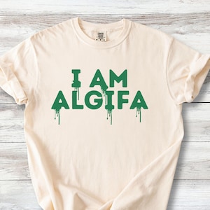 Könnte beinhalten: Ein cremefarbenes Kurzarm-T-Shirt mit dem Text I AM ALGIFA in dunkelgrüner Schrift. Die Buchstaben haben einen markanten Tropfeffekt, bei dem vertikale Farbspuren von der Unterseite jedes Buchstabens nach unten verlaufen.