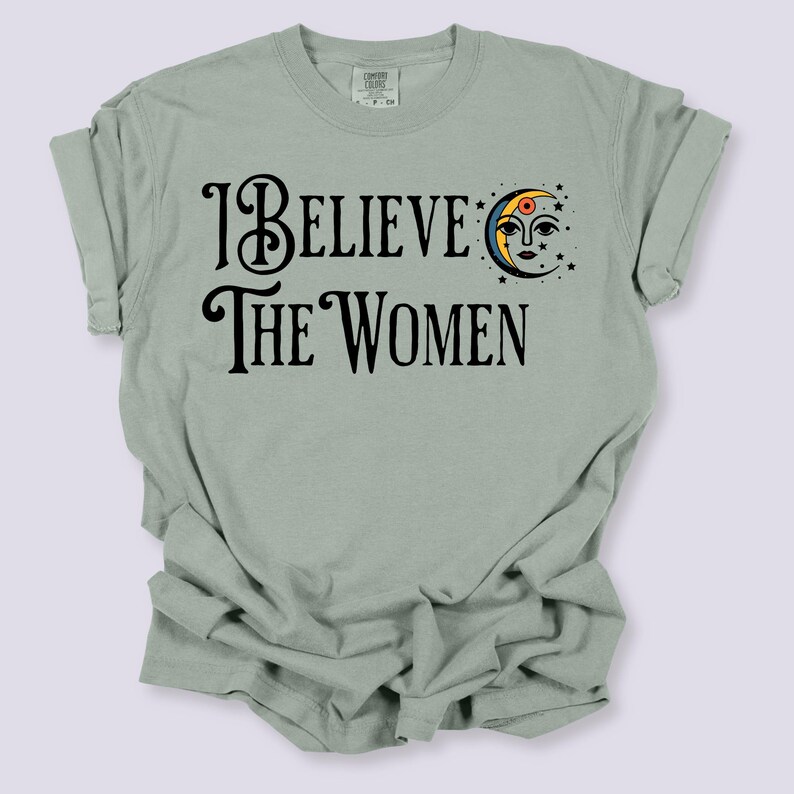 May include: Sage green t-shirt with the text "I Believe The Women" in black script. A crescent moon with a face and stars is on the right side of the text. The shirt has short sleeves and a crew neck.