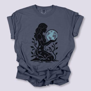 May include: A blue-grey t-shirt features a black graphic of a woman holding a globe, surrounded by leaves. The text "Reclaim Mother Earth" is arched above the globe. The shirt has rolled-up sleeves and a crew neck.