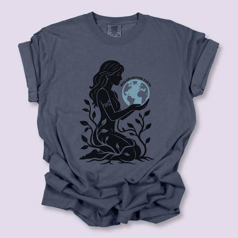 May include: A blue-grey t-shirt features a black graphic of a woman holding a globe, surrounded by leaves. The text "Reclaim Mother Earth" is arched above the globe. The shirt has rolled-up sleeves and a crew neck.