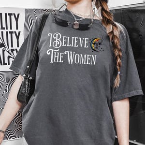 May include: Dark gray t-shirt with the text "I Believe The Women" in white script. A crescent moon and sun graphic is on the right side. The shirt has short sleeves and a relaxed fit.