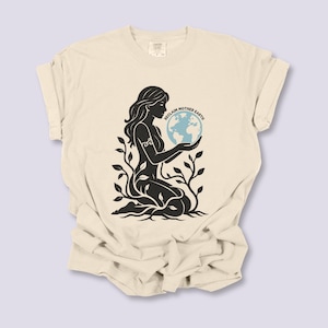 May include: Cream-colored t-shirt featuring a black silhouette of a woman holding a blue globe, with the text "RECLAIM MOTHER EARTH". The woman is surrounded by plant-like imagery.