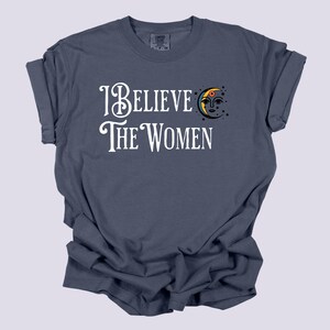 May include: A navy blue t-shirt with the text "I Believe The Women" in white script. A crescent moon graphic with a face and stars is to the right of the text. The shirt is made of a soft, comfortable material.