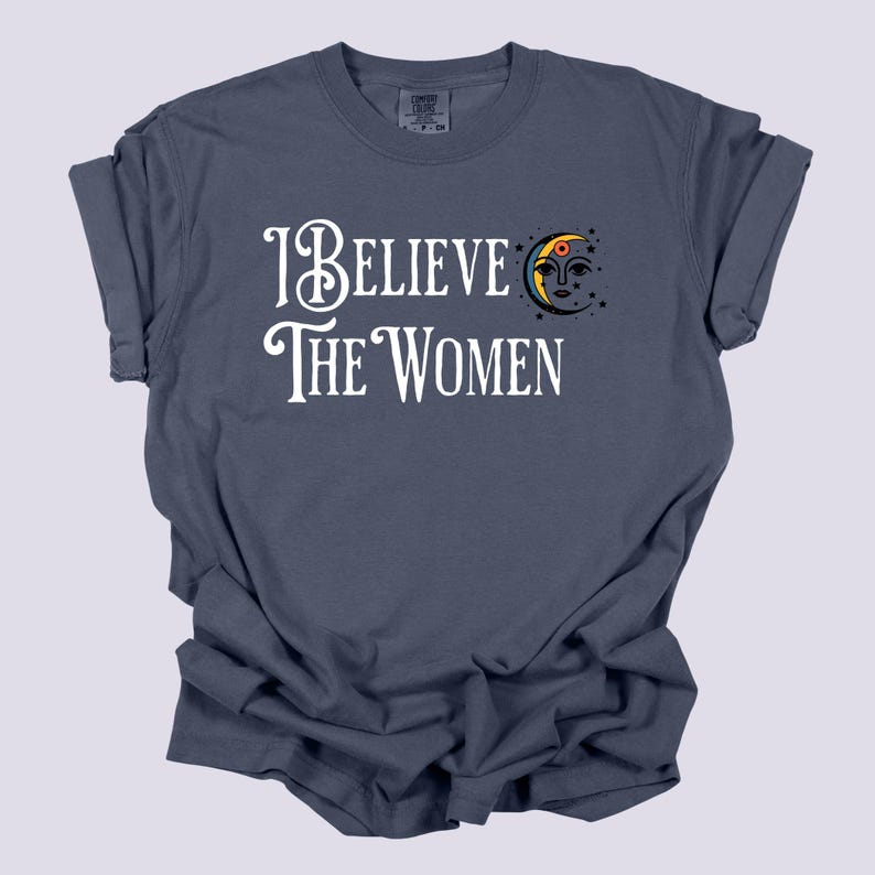 May include: A navy blue t-shirt with the text "I Believe The Women" in white script. A crescent moon graphic with a face and stars is to the right of the text. The shirt is made of a soft, comfortable material.