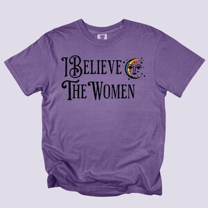 May include: A purple t-shirt with the text "I Believe The Women" in black script. A crescent moon with a face and stars is above the word "Women". The shirt is a solid color and has short sleeves.