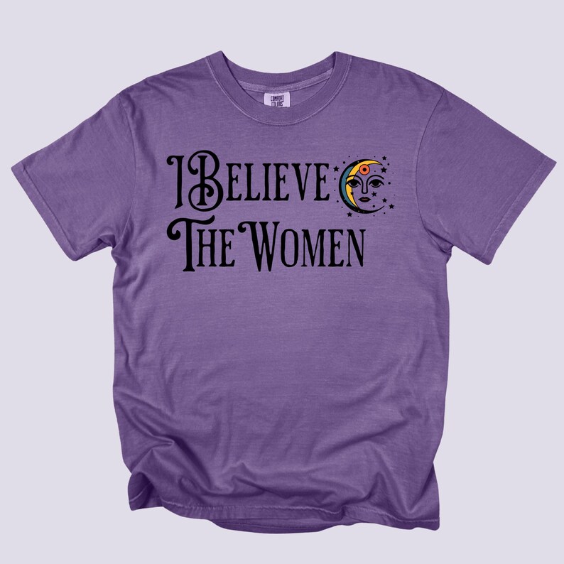 May include: A purple t-shirt with the text "I Believe The Women" in black script. A crescent moon with a face and stars is above the word "Women". The shirt is a solid color and has short sleeves.
