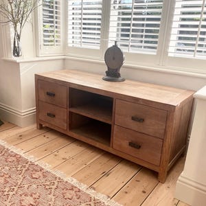 May include: A rectangular wooden media console featuring four drawers with dark metal handles and two open central shelves. The unit has a natural light brown wood grain finish and sits on a flat base. A dark metal scale sits on the top surface.