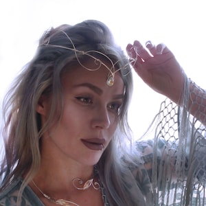 May include: A woman wearing a silver-toned headpiece and necklace with clear gemstone accents. The headpiece is delicate and sits atop her light-coloured hair. The necklace has a flowing design. She is wearing a teal-coloured top.