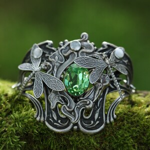 May include: A silver bracelet featuring a green gemstone and two dragonfly accents. The bracelet is resting on a bed of green moss.