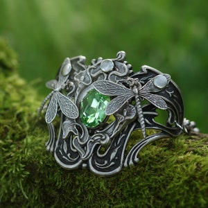 May include: A silver bracelet featuring a green gemstone and dragonfly accents. The bracelet is intricately designed with floral motifs and has a delicate chain.