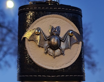 May include: A black leather flask with a silver bat design on a white circle. The flask has a silver cap and a black stitched border.