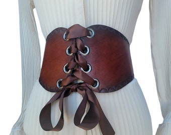 May include: A brown leather corset belt with a lace-up front and brown satin ribbon ties. The belt has silver eyelets and is worn over a white blouse.
