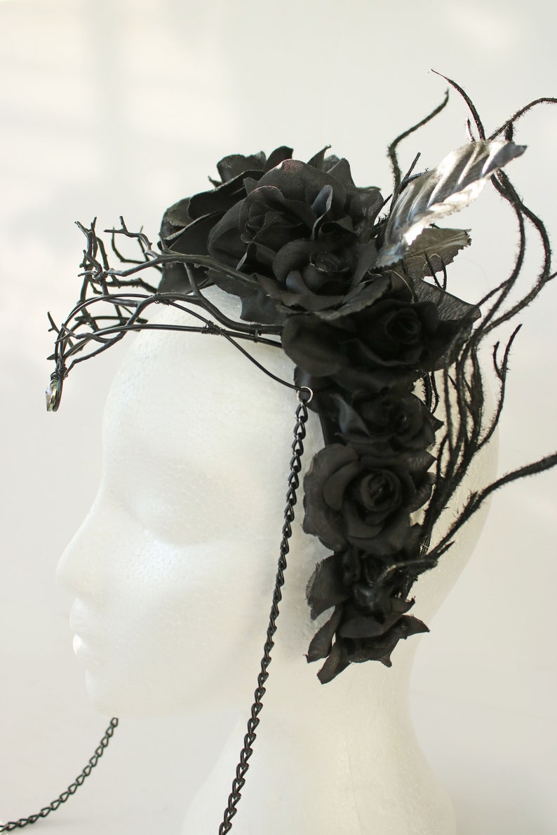 May include: A black floral crown with black roses and branches, accented with a black chain and a single black feather.