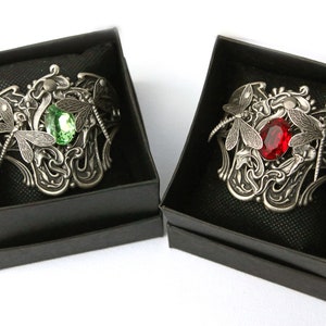 May include: Two silver bracelets with dragonfly designs and jeweled accents. One bracelet features a green gemstone, while the other has a red gemstone.