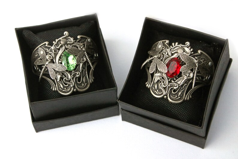 May include: Two silver bracelets with dragonfly designs and jeweled accents. One bracelet features a green gemstone, while the other has a red gemstone.