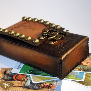 May include: A brown leather box with a brass clasp and studs. The box is closed and sitting on top of several tarot cards. The cards are face up and show images of people and symbols.