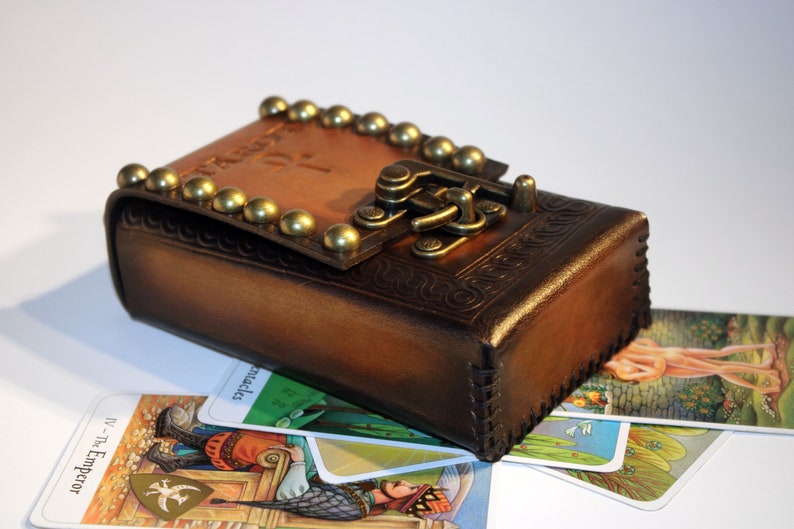 May include: A brown leather box with a brass clasp and studs. The box is closed and sitting on top of several tarot cards. The cards are face up and show images of people and symbols.