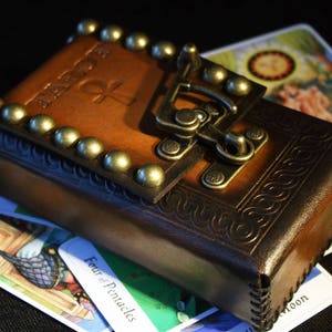 May include: A brown leather tarot card deck box with a brass clasp and decorative studs. The box is closed and has the word "Tarot" embossed on the front.